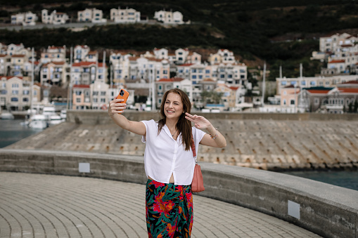 Smiling young woman using a smartphone and taking selfie during her summer vacation at the seaside in Lustica Bay