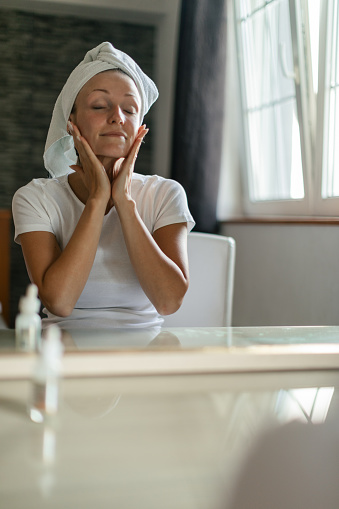 Satisfied young woman take care of her skin with hair wrapped in a towel, after showering. She is applying lotion, face milk and moisturizer in front of mirror in bathroom. She looks beautiful and fresh