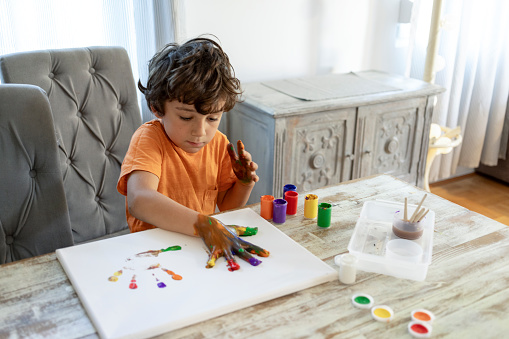 The preschooler, a six year-old with curly hair, eagerly starts his day by drawing vibrant, colorful pictures with watercolors on canvas in the cozy living room of his home each morning. At home in the morning, a seven-year-old boy with curly hair enthusiastically tackles his homework and unleashes his creativity by using water temperas of various colors to paint on canvas in the welcoming living room.