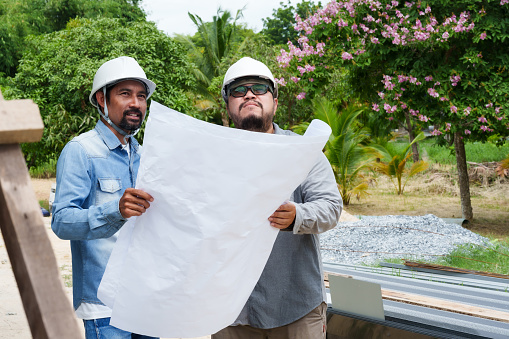 Two engineer men wearing safety helmets for safety while in construction site, hold large sheet house plan paper in hand check completeness according customer's needs, talk with casual expressions.
