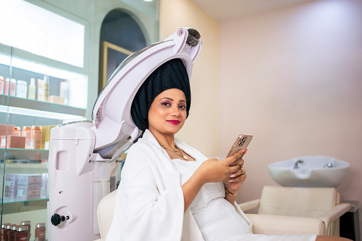 Young woman using her smartphone and chatting while having hair treatment in salon.