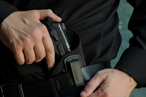 A man in black clothes takes out a black pistol from the holster, which is located on the waist belt of his trousers.