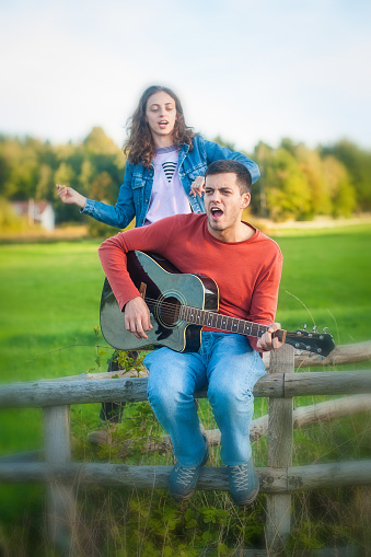 Romantic happy cute caucasian couple playing guitar and singing songs sitting on wooden fence in meadow in outdoor nature park