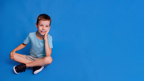 schoolboy hold his palm to cheeks, thinking and wearing blue mockup T-shirt over light blue studio background