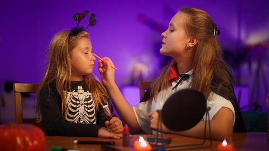 A mother is putting on Halloween make up on her daughter's face in the living room at home.