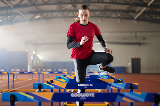 Young male runner training and jumping through hurdles at indoors stadium running track. Sport and competition concept.