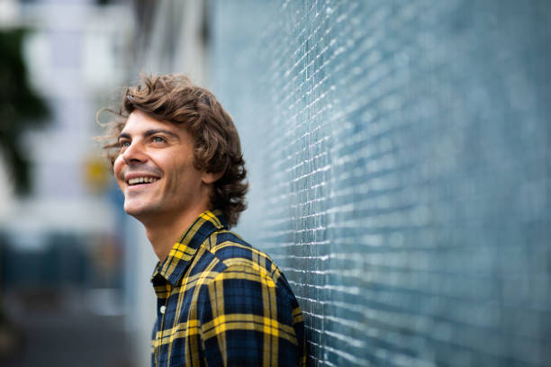 Smiling young man leaning against gray wall and looking up