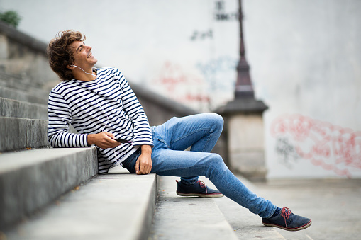 Portrait of happy young man relaxing on steps outside listening to music with earphones and cellphone