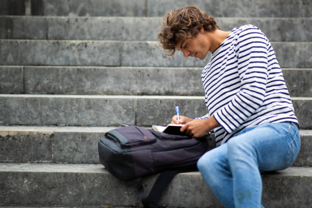 istock Young male student sitting on steps outside and writing in book 1678482870
