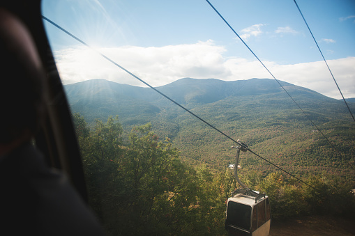 The view from inside a gondola or ski lift or chair lift going up the mountain side. Mount Washington is in view with lots of tree and sunlight coming through.
