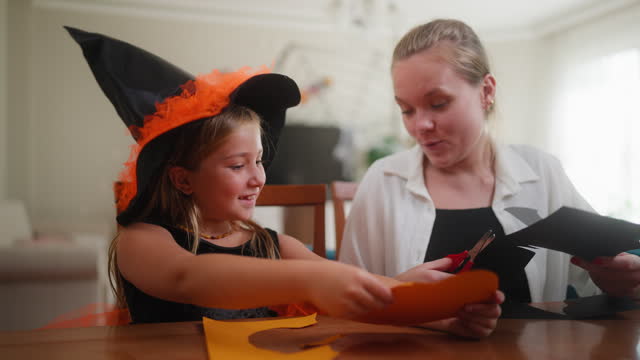Small girl making Halloween decorations with her mother in living room at home