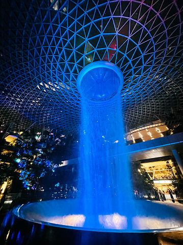 Jewel Changi Airport, Singapore - 11th October, 2022: Low angle view of illuminated Rain Vortex inside the Jewel area at Changi Airport at night. It's the world's tallest waterfall at 130 ft in height and surrounded by a four-storey terraced forest. The Jewel complex and waterfall was designed by Moshe Safdie.