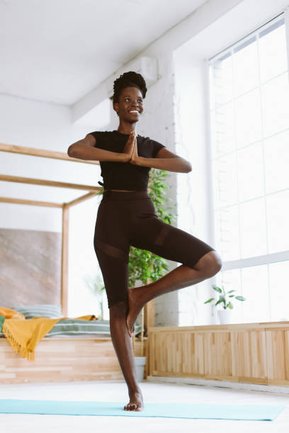 istock Vertical smiling African woman with dreadlocks practicing yoga, tree pose asana on mat at home. Low angle view 1678545565