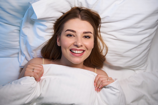 Top view of smiling female covered with blanket looking at camera while lying on bed indoors