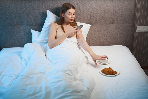 Caucasian female in bra using mobile while lying in bed with breakfast indoors