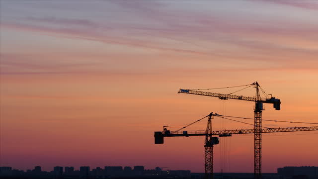 Time lapse. Silhouettes of working cranes on the red colored sky background at the sunset time. Workers are doing something at the top of one crane