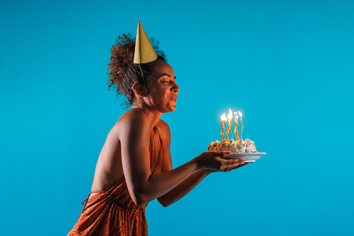 Profile view of happy young woman, in a party hat, standing against blue background and blowing out candles on a birthday cake that she is holding.