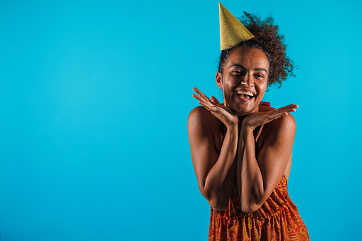Portrait of cheerful young woman in a party hat, standing against blue background, framing her face with hands and joyfully smiling at camera.