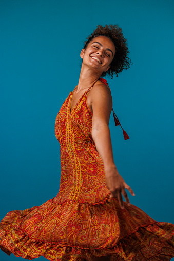 Portrait of cheerful young woman dancing in a flowy orange colored dress, against blue background, looking at camera and joyfully smiling.