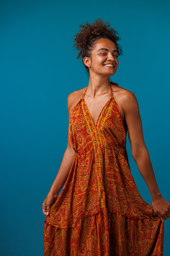 Portrait of cheerful young woman dancing in a flowy orange colored dress, against blue background, looking at camera and joyfully smiling.