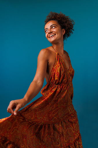 Portrait of cheerful young woman dancing in a flowy orange colored dress, against blue background, looking at camera and joyfully smiling.