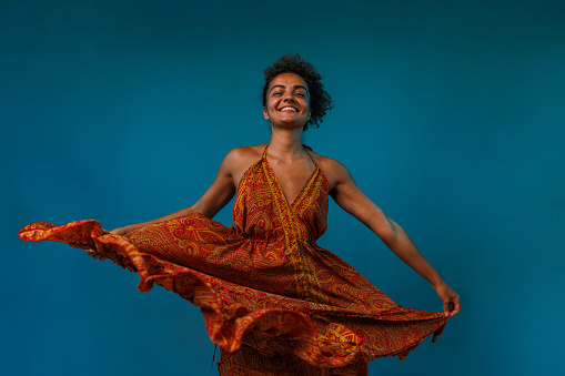 Portrait of cheerful young woman dancing in a flowy orange colored dress, against blue background, looking at camera and joyfully smiling.