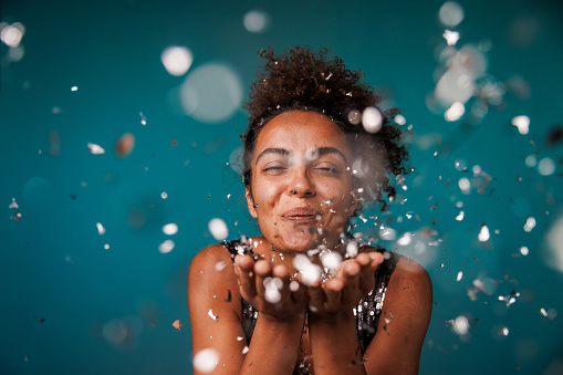 Candid shot of cheerful young woman standing against dark teal blue background and blowing silver colored confetti at camera.