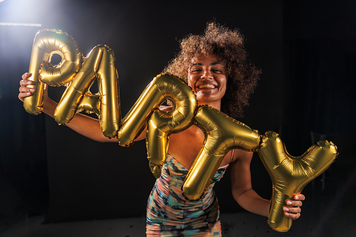 Portrait of cool young woman dancing against black background, holding gold colored balloons that spell the word ''party'' and cheerfully smiling at camera.