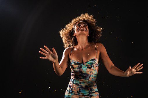 Portrait of cool young woman with curly hair dancing against black background while gold colored confetti are exploding behind her.