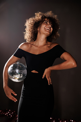 Portrait of cool young woman in black outfit standing against dark gray background, hand on hip, holding a disco ball.