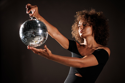 Portrait of cool young woman in black outfit standing against dark gray background and playing with a disco ball that she is holding.