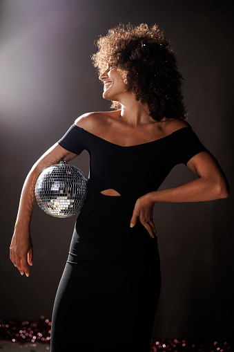 Portrait of cool young woman in black outfit standing against dark gray background and playing with a disco ball that she is holding.