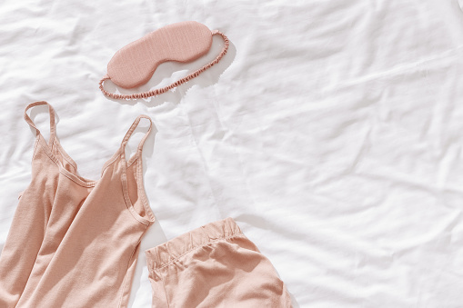 Top view beige pajama and eye mask on white bedcloth background, copy space. Cozy pyjamas for comfort rest at night. Flat lay of singlet, shorts, sleeping mask neutral light color, sleep well concept