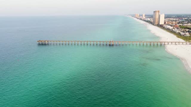 Aerial of Panama City Beach Pier at Pier Park over the gulf of mexico turquoise blue and green water. Aerial shot of the silhouette of a fishing pier on the ocean with a colorful and golden sunrise.