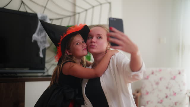 Young mother taking selfies with her small daughter dressed in Halloween witch costume in living room at home