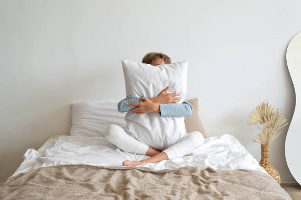 time to sleep, sleep deficiency, good morning concept. teenager boy sitting on bed, embracing white pillow, hiding face behind. minimalist scandinavian neutral bedroom interior, lifestyle - one person child serene people failure imagens e fotografias de stock