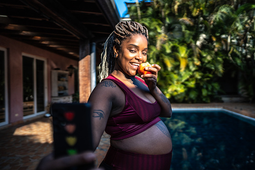 Pregnant woman taking a selfie using mobile phone eating an apple after exercising on home workout