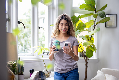 Happy woman looking at social media on her cell phone while drinking a cup of coffee at home - lifestyle concepts