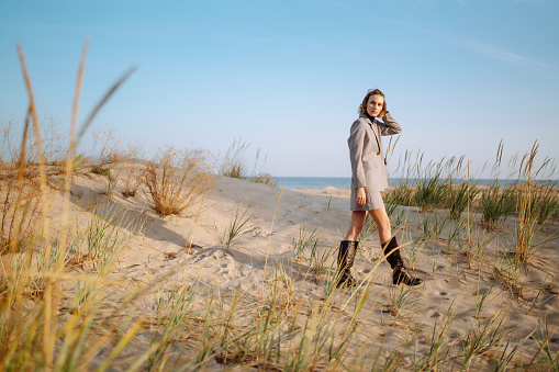 Stylish woman posing on autumn beach in wild grass. A woman enjoys the autumn nature on the beach, relaxing. Fashion, style and relaxation concept.