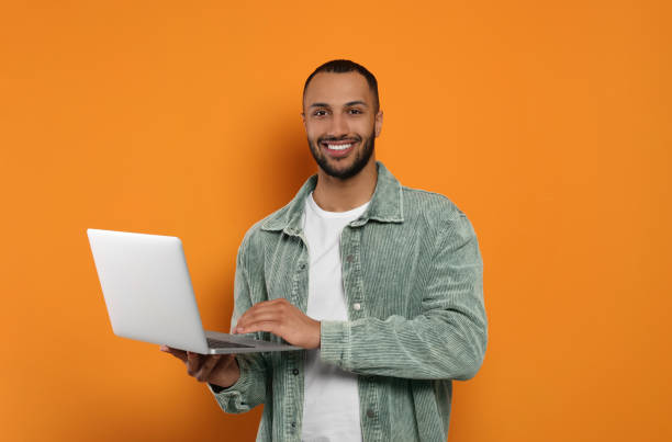 istock Smiling young man working with laptop on orange background 1678874356