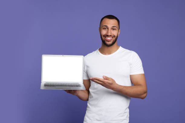 istock Smiling young man showing laptop on lilac background 1678874842