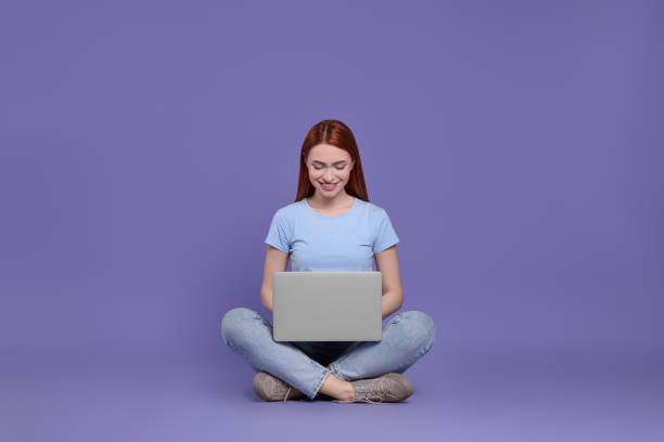 istock Smiling young woman working with laptop on lilac background 1678893353