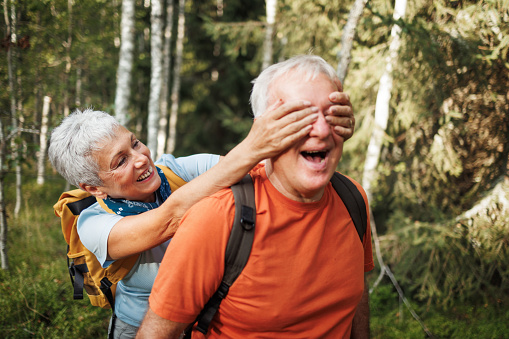 Senior couple having fun while hiking together on a sunny day, wife is covering her husbands eyes with hands from behind