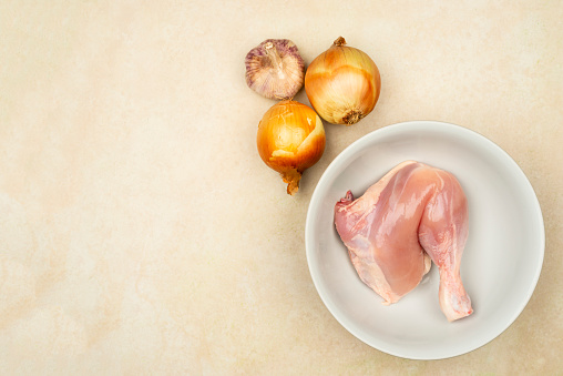 High angle view of of raw chicken thigh in a white plate with ingredients to prepare as onion and garlic