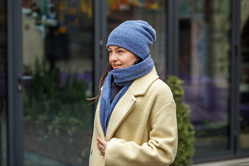 Stylish attractive young smiling woman walking in street in winter outfit wearing coat, blue knitted hat and scarf, happy mood, fashion style trend