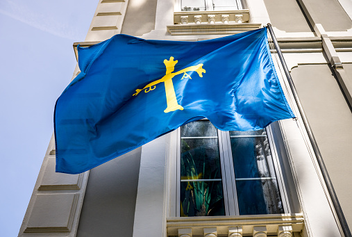 Flag of Asturias of Blue Color with the Yellow Cross Waving in a Garden with Gray Building in the Background. View from below