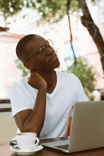 Vertical african american, dark skin man in glasses, sitting with laptop in street cafe, touching and massaging his neck, feeling uncomfortable pain after long sedentary work. Problem of wrong posture