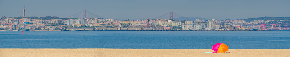 panoramic view of downtown Lisbon seen from the river beach of Barreiro with a tongue of white sand with a colorful parasol in a minimalist representation. Tagus River between the two cities.  The statue of Christ the King of Almada and the 25 de Abril bridge are visible