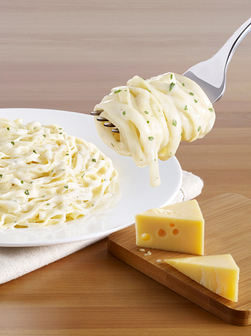Forkful of delicious creamy pasta in front of a pasta plate and cheese blocks on a wooden table