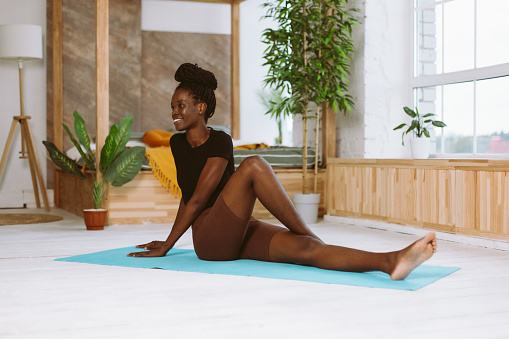 Smiling fit African woman turning body sitting on floor with bent leg, training on gymnastic mat in decorated studio. Muscle stretching, gymnastic exercises, fitness, keeping body fit.